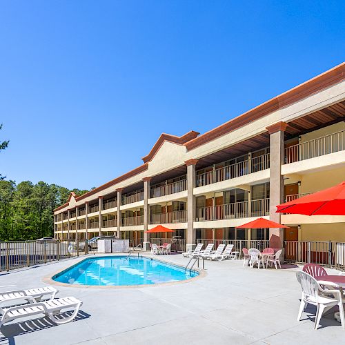 A sunny motel-style pool area with a long two-story beige building, red umbrellas, lounge chairs, and a bright blue pool.