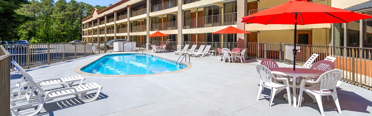 A hotel pool area with lounge chairs, red umbrellas, and tables, surrounded by a multi-story building and trees.