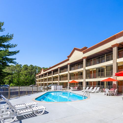 A hotel pool area with lounge chairs, red umbrellas, and tables, surrounded by a multi-story building and trees.