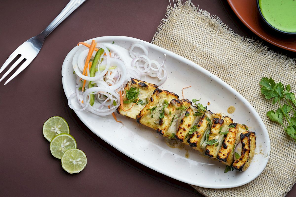 Grilled paneer slices garnished with herbs, served with sliced onions, carrots, and limes on a white plate with a green dip and fork.