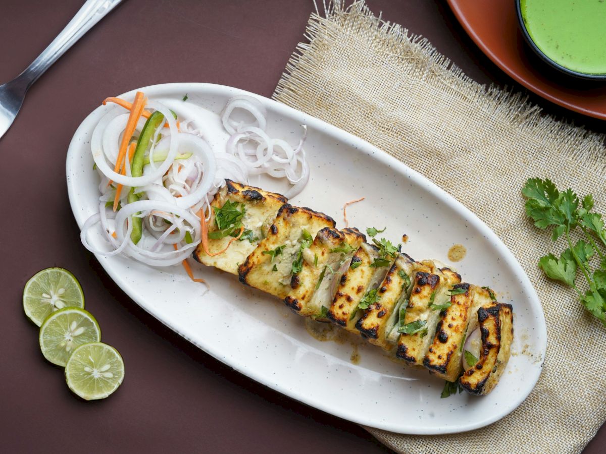 Grilled paneer slices garnished with herbs, served with sliced onions, carrots, and limes on a white plate with a green dip and fork.