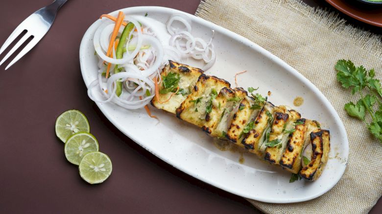 Grilled paneer slices garnished with herbs, served with sliced onions, carrots, and limes on a white plate with a green dip and fork.