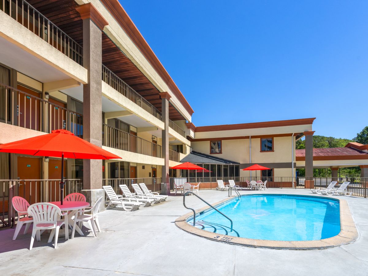 A motel pool area with a kidney-shaped blue pool, white lounge chairs, red umbrellas, and a tan multi-story building in sunny weather.