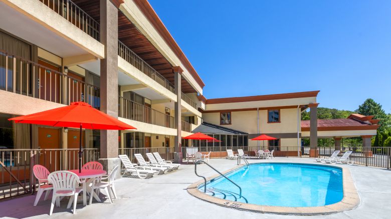 A motel pool area with a kidney-shaped blue pool, white lounge chairs, red umbrellas, and a tan multi-story building in sunny weather.