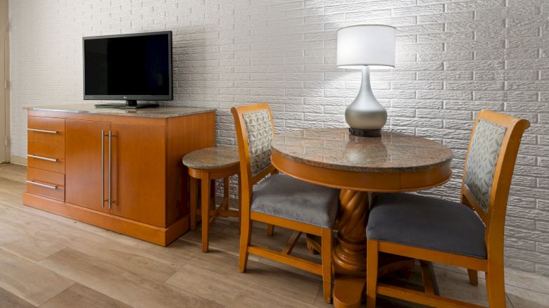 A small dining area with a round wooden table and two upholstered chairs, a wooden sideboard with a TV on top, and a lamp on the table.