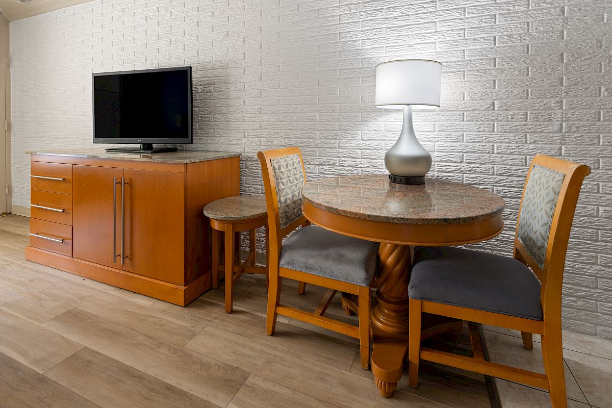 The image shows a room with a TV on a wooden cabinet, a round table with chairs, and a lamp against a white brick wall.
