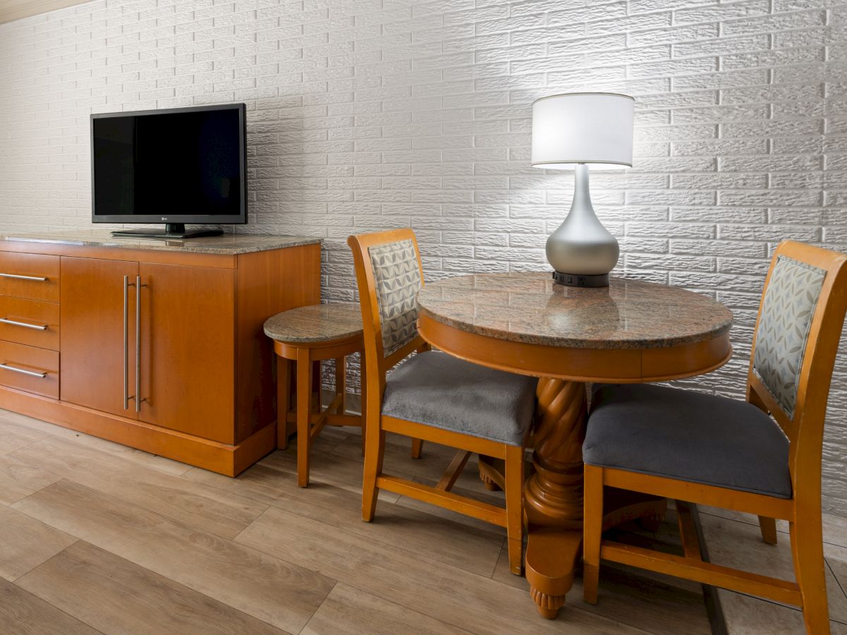 The image shows a room with a TV on a wooden cabinet, a round table with chairs, and a lamp against a white brick wall.