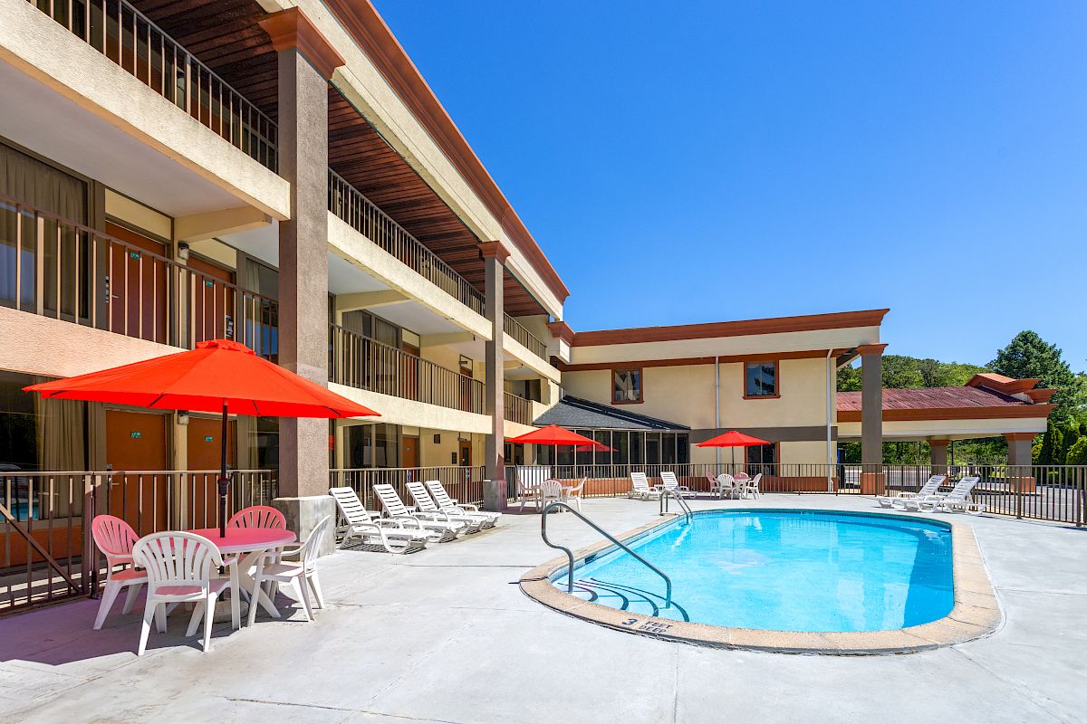 A hotel courtyard features a swimming pool, red umbrellas, lounge chairs, tables, and a three-story building under a clear blue sky.