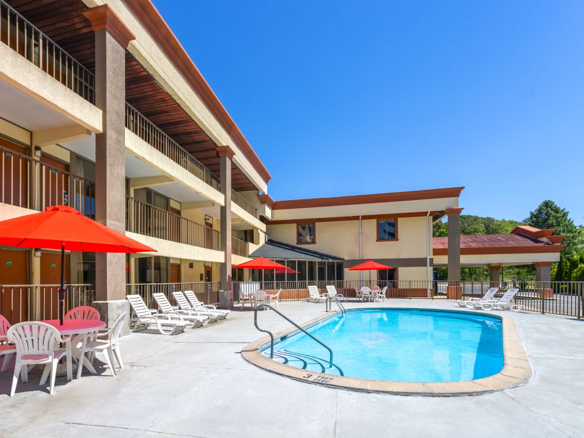 A hotel courtyard features a swimming pool, red umbrellas, lounge chairs, tables, and a three-story building under a clear blue sky.