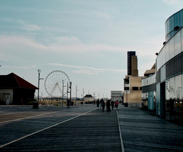 A boardwalk with a ferris wheel in the distance, modern buildings lining the right, a few people strolling, and a clear sky over the seaside.