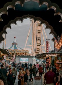 A bustling carnival scene viewed through an arch, featuring crowds, a carousel, and a Ferris wheel against a cloudy sky.