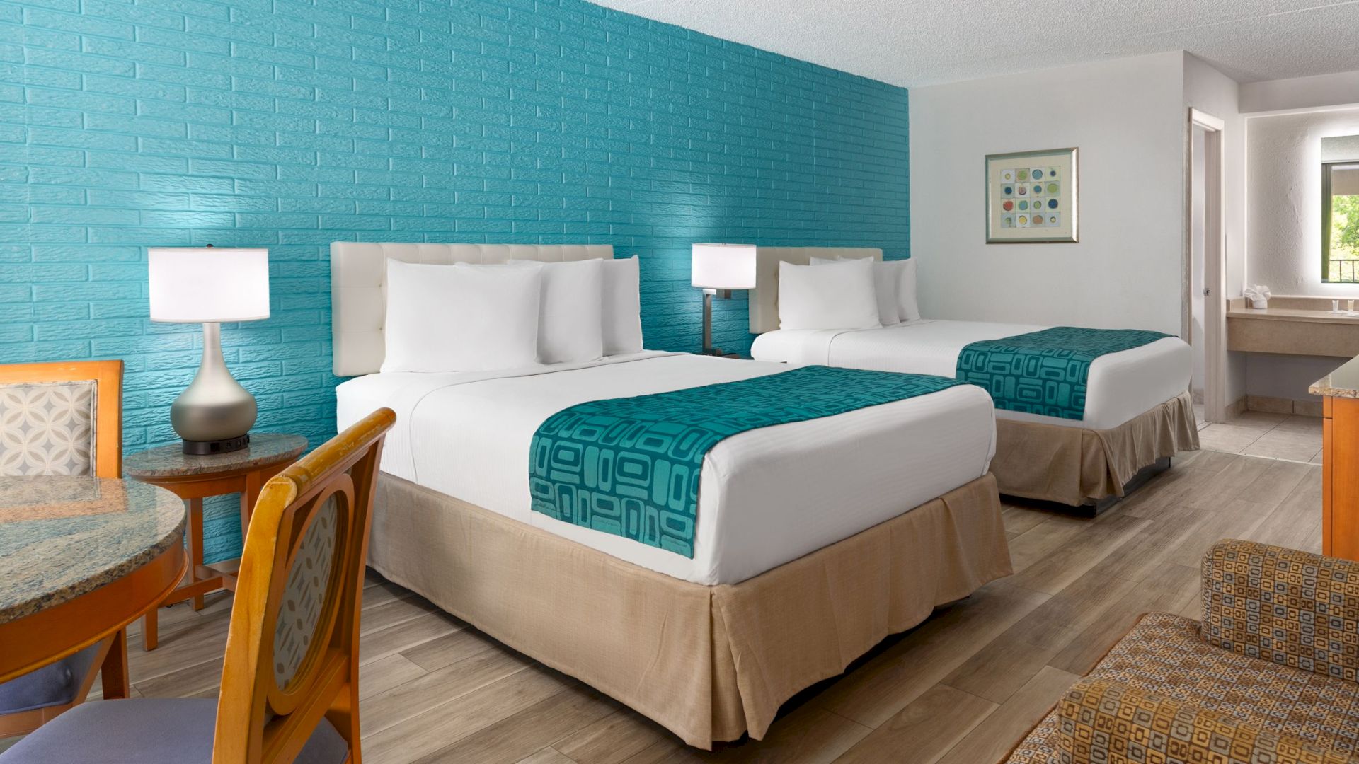 A hotel room with two beds, teal accent wall, nightstands with lamps, a small table and chairs, and a seating area across from a TV. (End.)