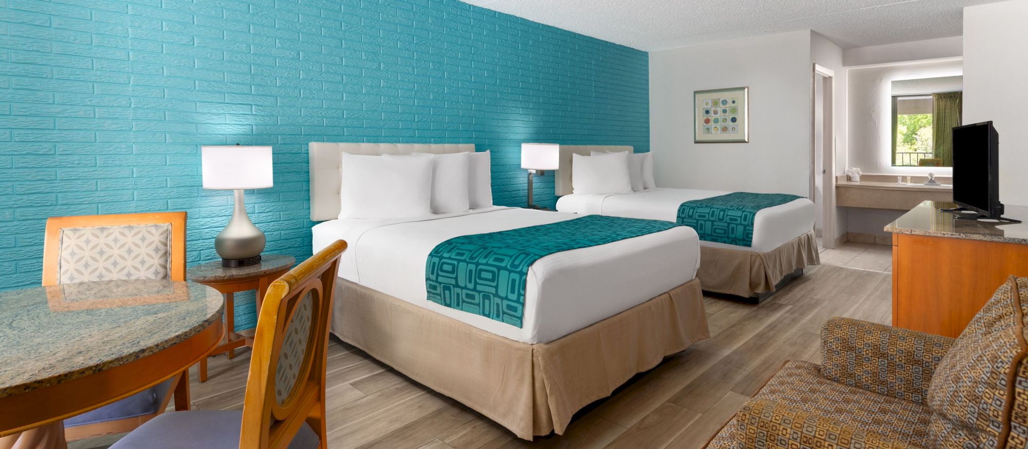 A hotel room with two beds, teal accent wall, nightstands with lamps, a small table and chairs, and a seating area across from a TV. (End.)