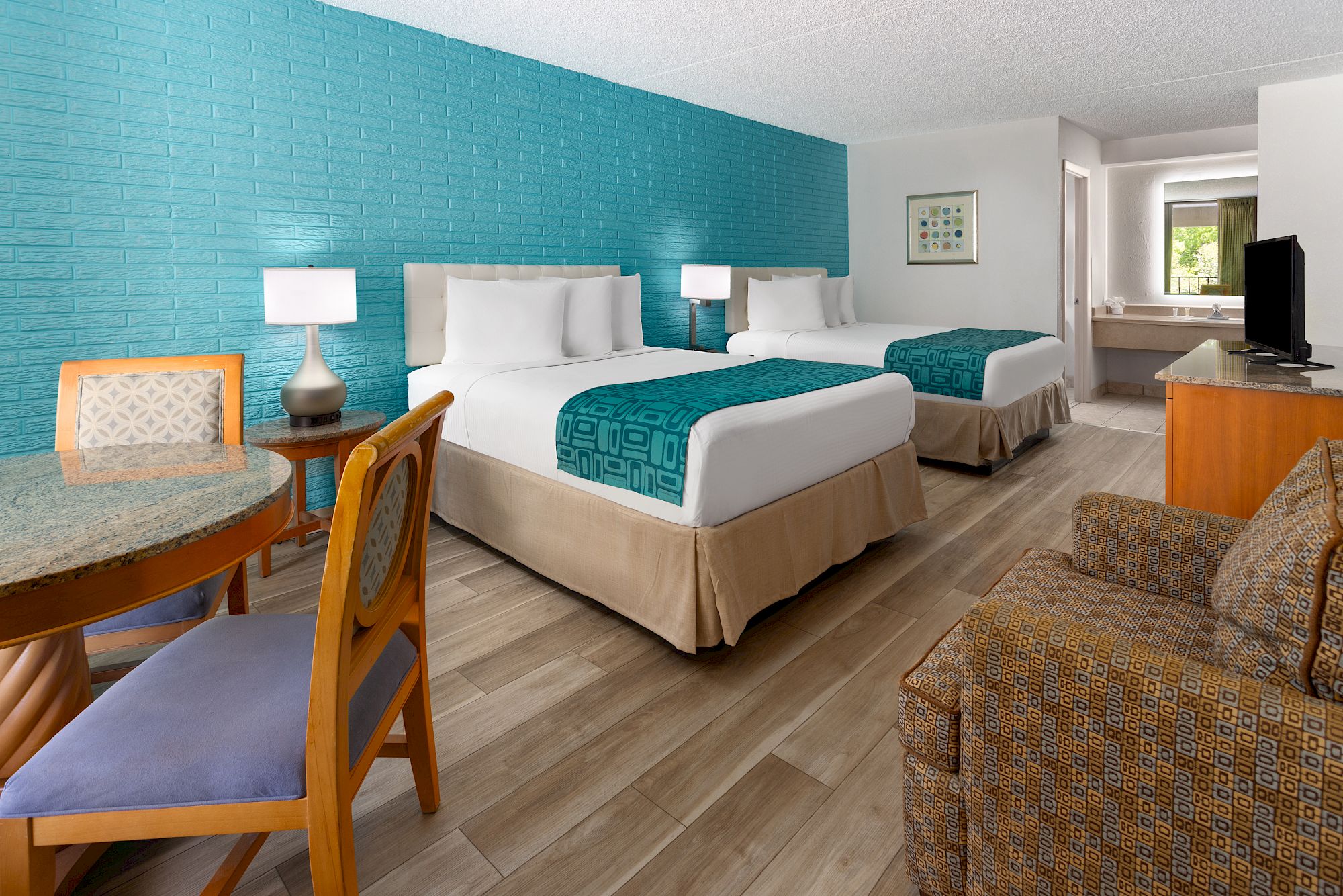 A hotel room with two beds, turquoise accents, a table with chairs, an armchair, and a TV. There's a bathroom area in the background.