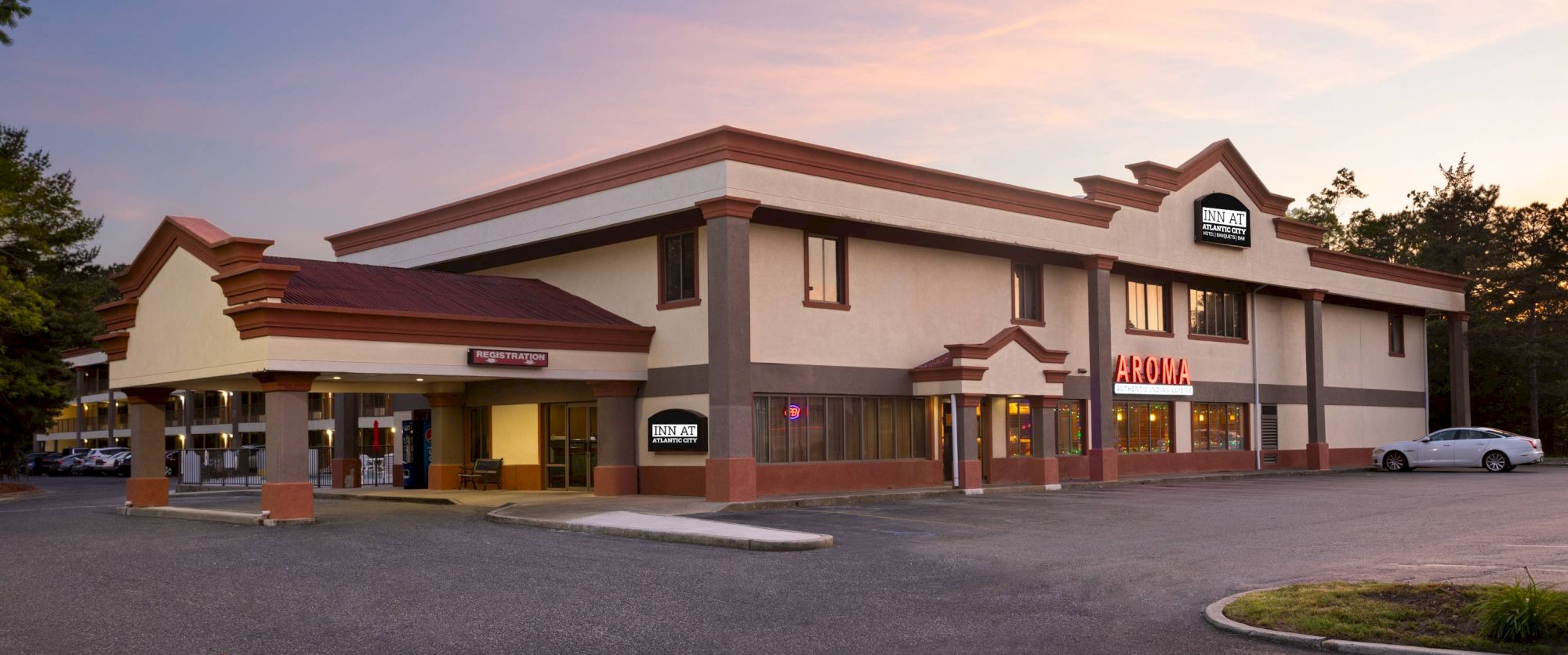 A white and tan two-story strip mall with brown trim, storefronts including Aroma, parking lot, and a sunset sky above.