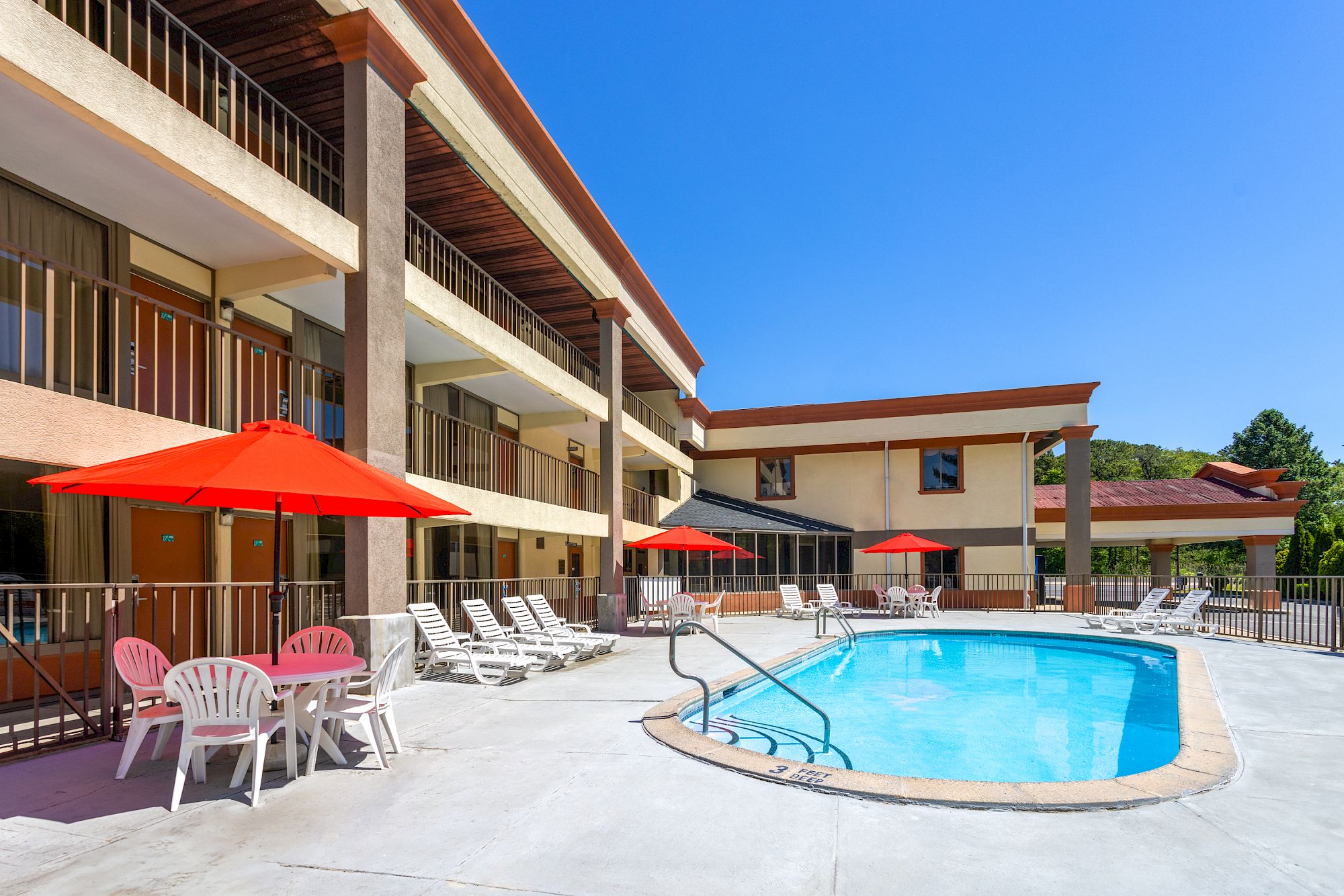 The image shows a hotel pool area with lounge chairs, red umbrellas, and a multi-story building in the background under a clear blue sky.