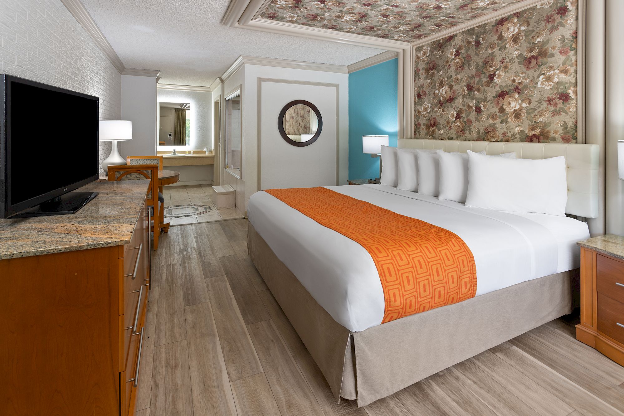 A hotel room with a large bed, orange throw, TV, wooden furniture, and a floral accent wall with a vanity area in the background.
