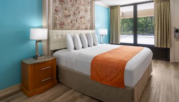 The image shows a hotel room with a neatly made bed, orange bed runner, wood flooring, teal walls, and a window with curtains.