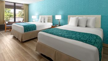 The image shows a hotel room with two double beds, turquoise decor, wooden flooring, bedside tables, and a view through the window.