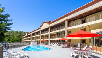 Outdoor pool area at a hotel with lounge chairs, red umbrellas, and surrounding trees under a clear blue sky.