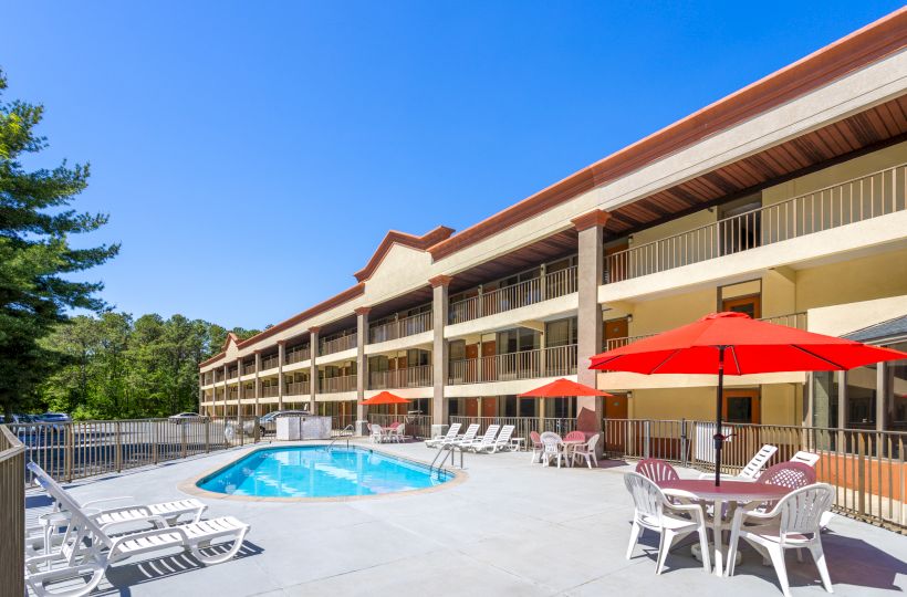 Outdoor pool area at a hotel with lounge chairs, red umbrellas, and surrounding trees under a clear blue sky.