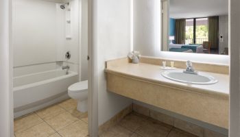 The image shows a bathroom with a bathtub, toilet, and sink area. A hotel room can be seen through an open doorway.