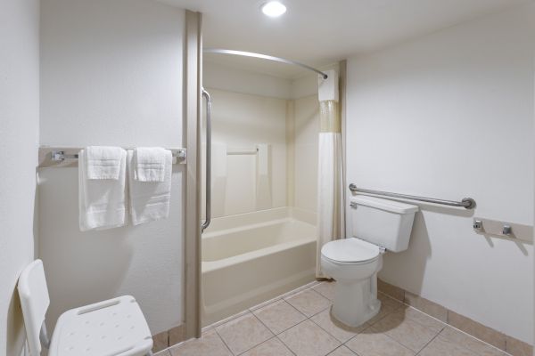 A bathroom featuring a shower-tub combo, a toilet with a grab bar, a shower chair, and towels hanging on a rail.