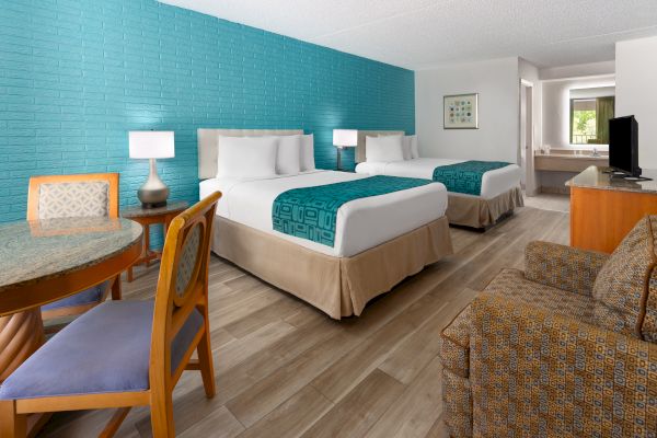 A hotel room with two beds, teal accent wall, table with chairs, TV, and a bathroom area. Cozy and modern design.