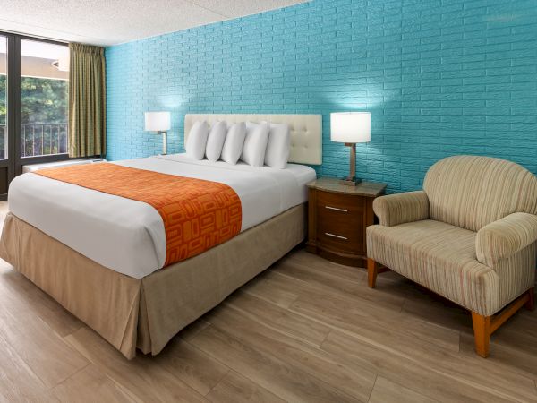 A hotel room with a bed, orange throw, two lamps, a bedside table, and an armchair against a blue wall, with a window view.