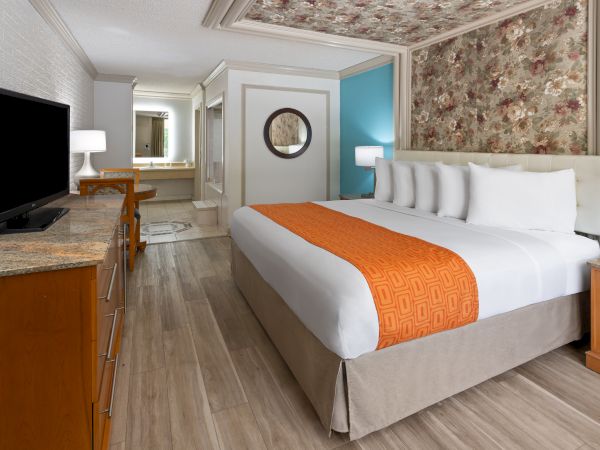A hotel room with a double bed, orange throw, TV, bedside table lamps, floral wall, and bathroom in the background.