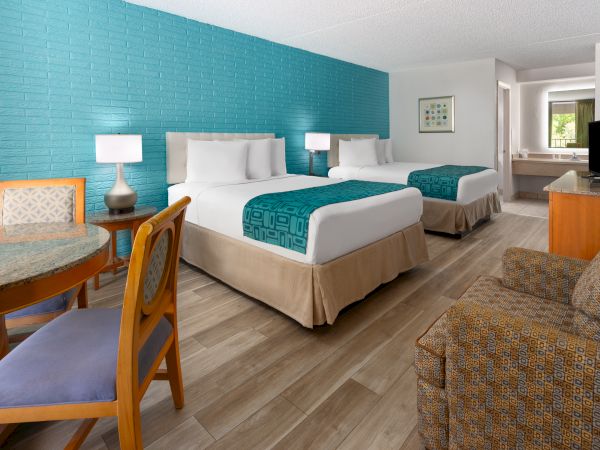 The image shows a hotel room with two beds, a turquoise accent wall, chairs, a small table, a sofa, lamps, a TV, and wood flooring.