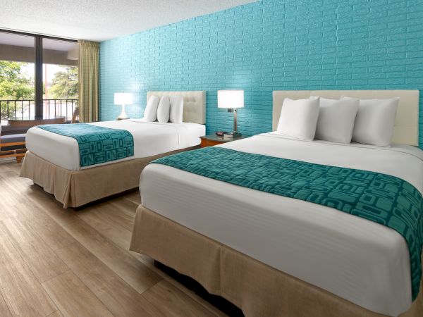 The image shows a hotel room with two beds, blue accent wall, wooden flooring, and a window with a view outside.