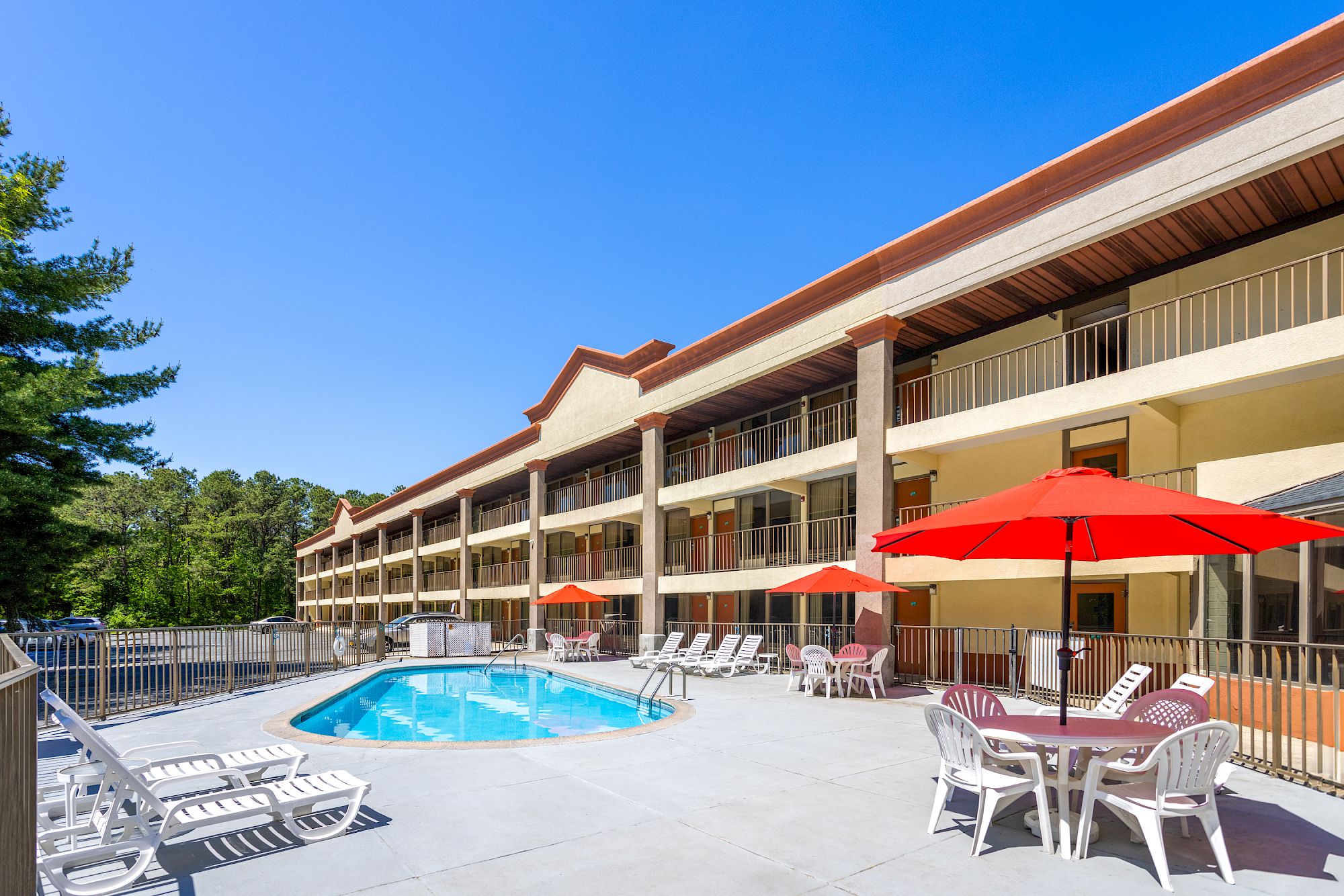 Outdoor hotel-style pool area with a multi-story building, lounge chairs, red umbrellas, and a bright blue sky.