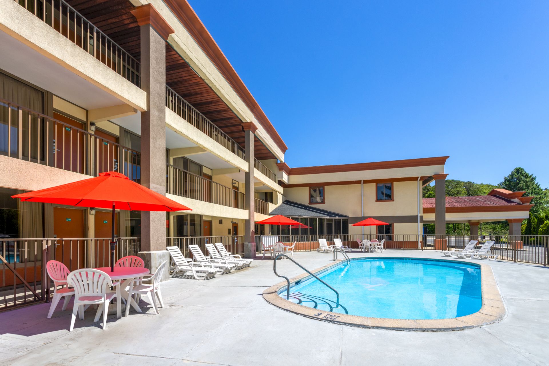 A sunny hotel pool area with a curved blue pool, red umbrellas, white lounge chairs, and a beige building with balconies overlooking the scene, ending with a period.