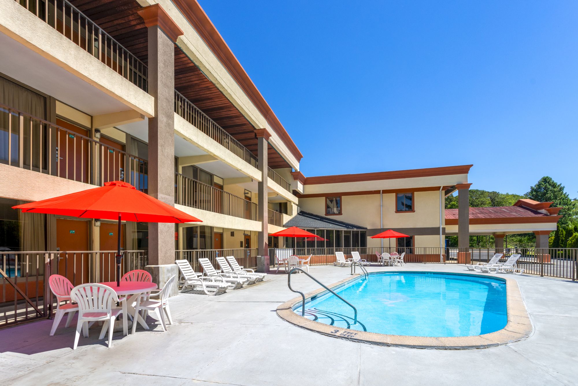 A sunny hotel pool area with a curved blue pool, red umbrellas, white lounge chairs, and a beige building with balconies overlooking the scene, ending with a period.