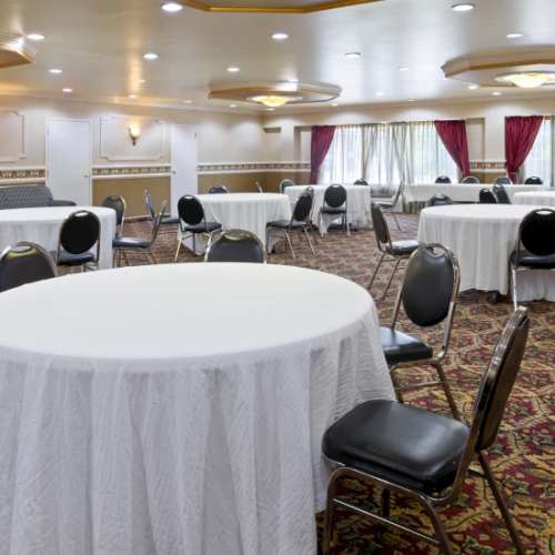 A bright banquet hall set with round tables, white linens, black chairs, and ceiling lights with red window curtains.