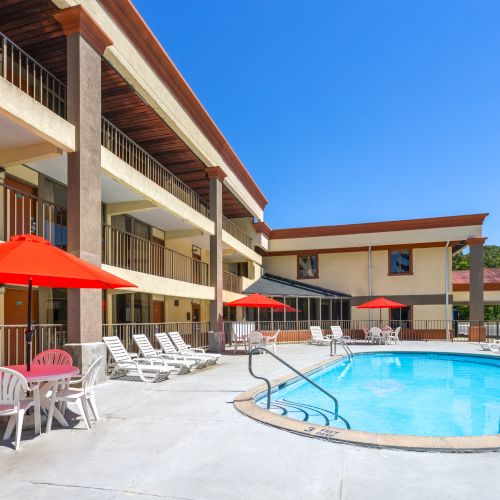 Outdoor hotel pool courtyard with blue water, red umbrellas, lounge chairs, and multi-story balconies under a clear sky.
