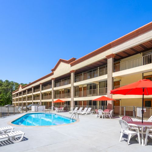 A sunny motel courtyard with a small outdoor pool, red umbrellas, white lounge chairs, and multi-story balconies against a clear blue sky.
