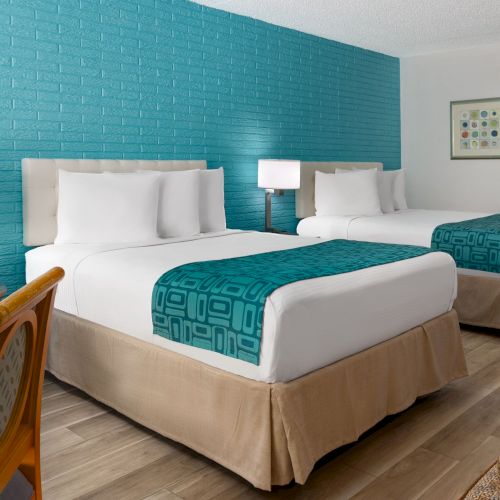 A bright hotel room with two white beds, teal accents, blue brick wall, nightstands, patterned chairs, and a TV by a dresser.
