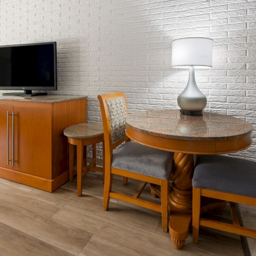 A cozy dining area with a round table and chairs, a TV on a wooden cabinet, and a floor lamp beside a white brick wall.