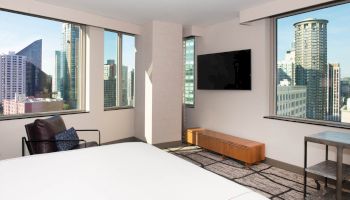 A modern hotel room with large windows showing a cityscape, a bed, TV, chair, and sleek furniture, creating a cozy, urban vibe.