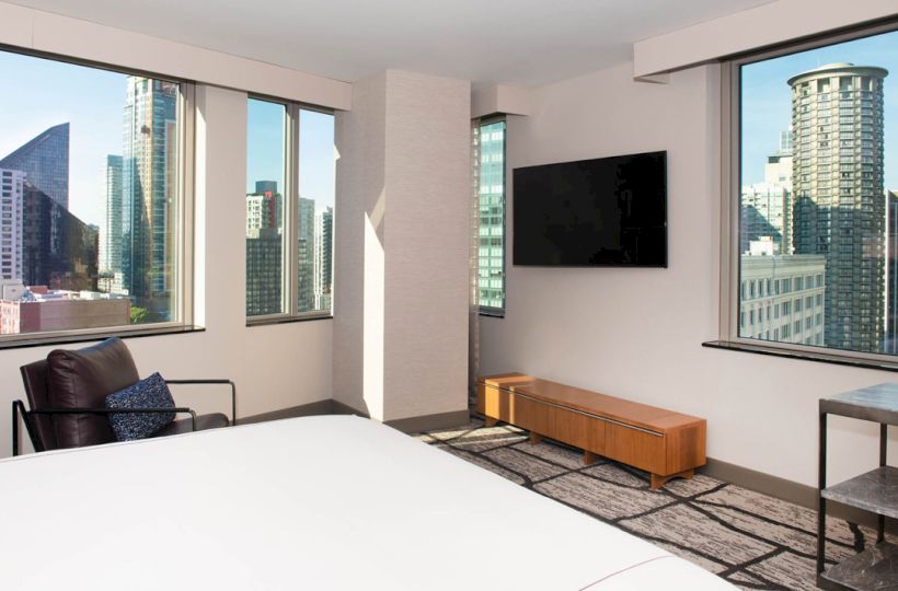 A modern hotel room with large windows showing a cityscape, a bed, TV, chair, and sleek furniture, creating a cozy, urban vibe.