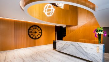 The image shows a modern lobby with a clock, marble reception desk, and spiral wood ceiling featuring decorative lights and flowers.
