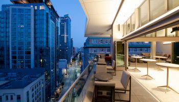 A modern balcony overlooks a cityscape with tall buildings at dusk, featuring tables and chairs for relaxation.