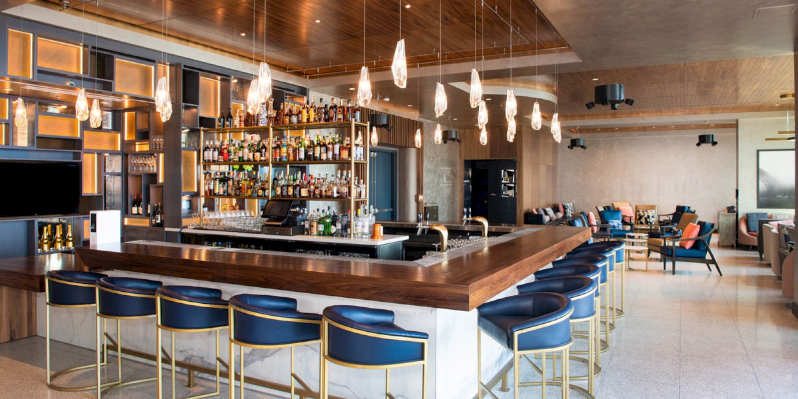 A modern bar with blue stools, a well-stocked bar, pendant lights, and seating area in the background, featuring a sleek design.