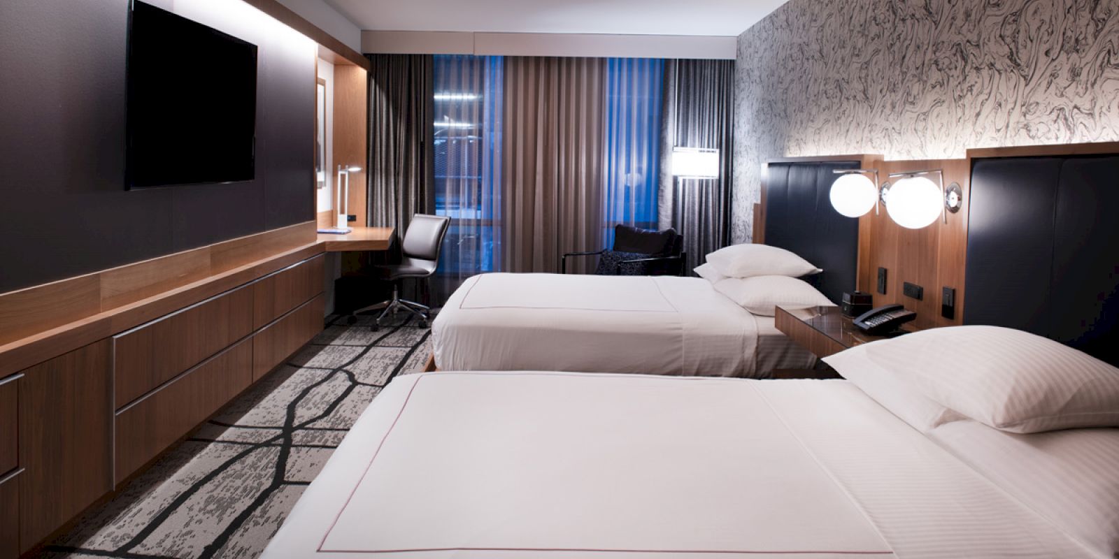 A modern hotel room with two beds, a flat-screen TV, desk, chair, and lamps, featuring a stylish wall design and large window.