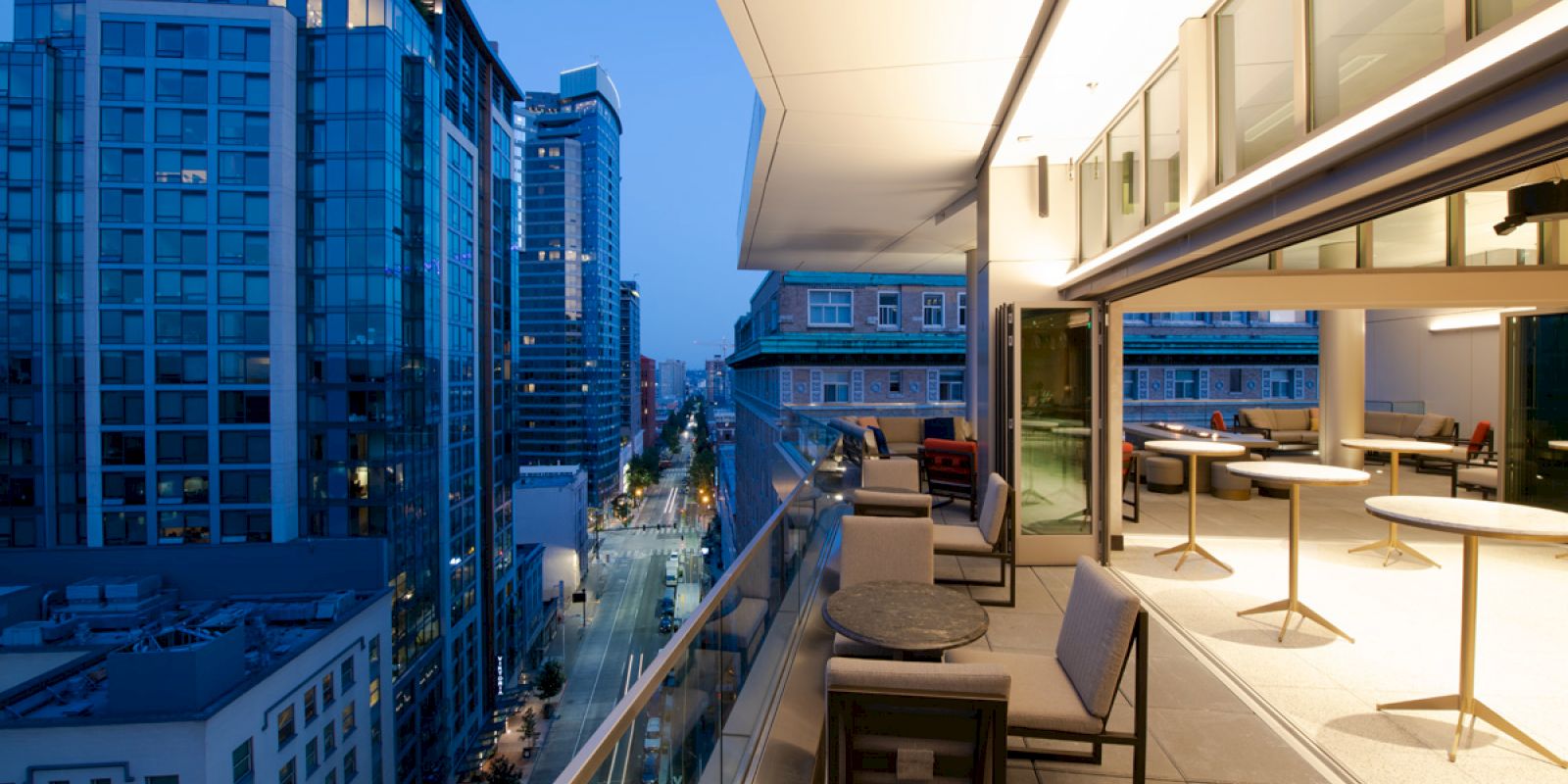 A modern balcony overlooking a cityscape with tall buildings at dusk, featuring outdoor seating and tables, illuminated warmly.