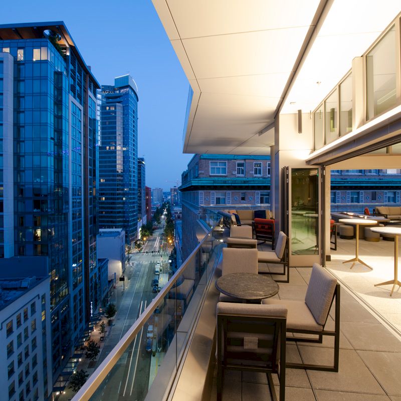 A modern balcony overlooks a city street with tall buildings, featuring outdoor seating and large windows, as evening approaches.