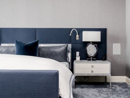 A modern bedroom with a blue headboard, white bedding, a nightstand with a lamp and decorative piece, and plush carpeting.
