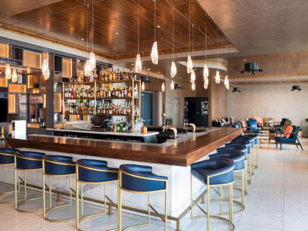 The image shows a modern bar with a central counter, barstools, hanging lights, and shelves filled with bottles.
